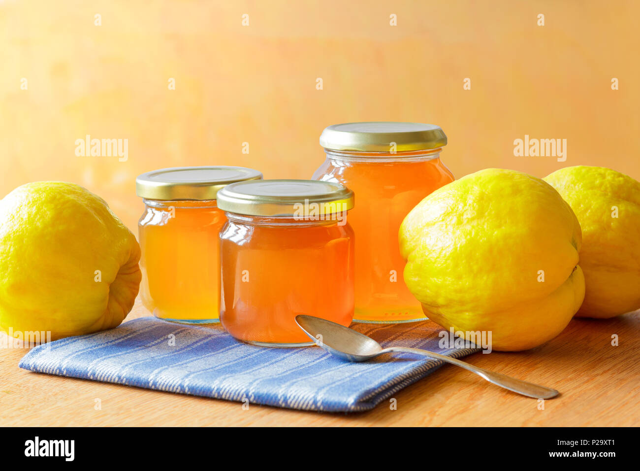 Quittengelee in Gläsern mit Quitten auf einem Holztisch in strahlendem Sonnenschein vor einem gelben und orangenen Hintergrund. Stockfoto