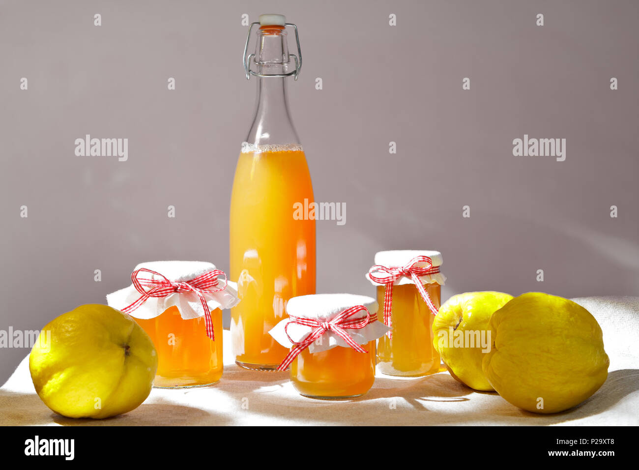 Hausgemachte Quitten Gelee und Saft in Flaschen und Gläsern mit Quitten auf Leinen Tischdecke im hellen Sonnenschein vor neutralem Hintergrund. Stockfoto