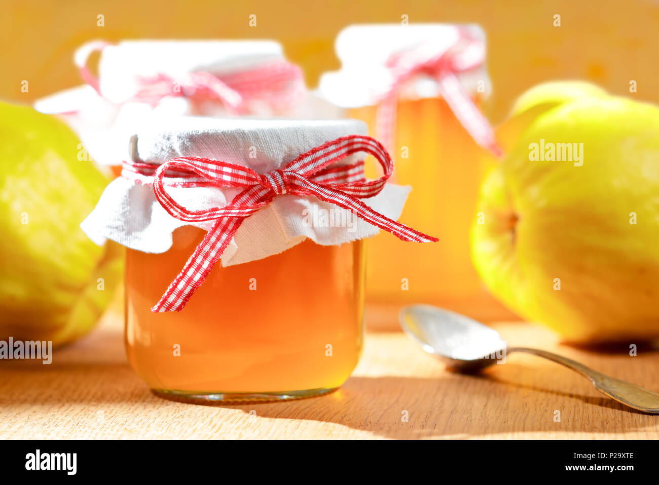 Quittengelee in Gläsern mit Quitten auf einem Holztisch in strahlendem Sonnenschein vor einem gelben und orangenen Hintergrund. Stockfoto