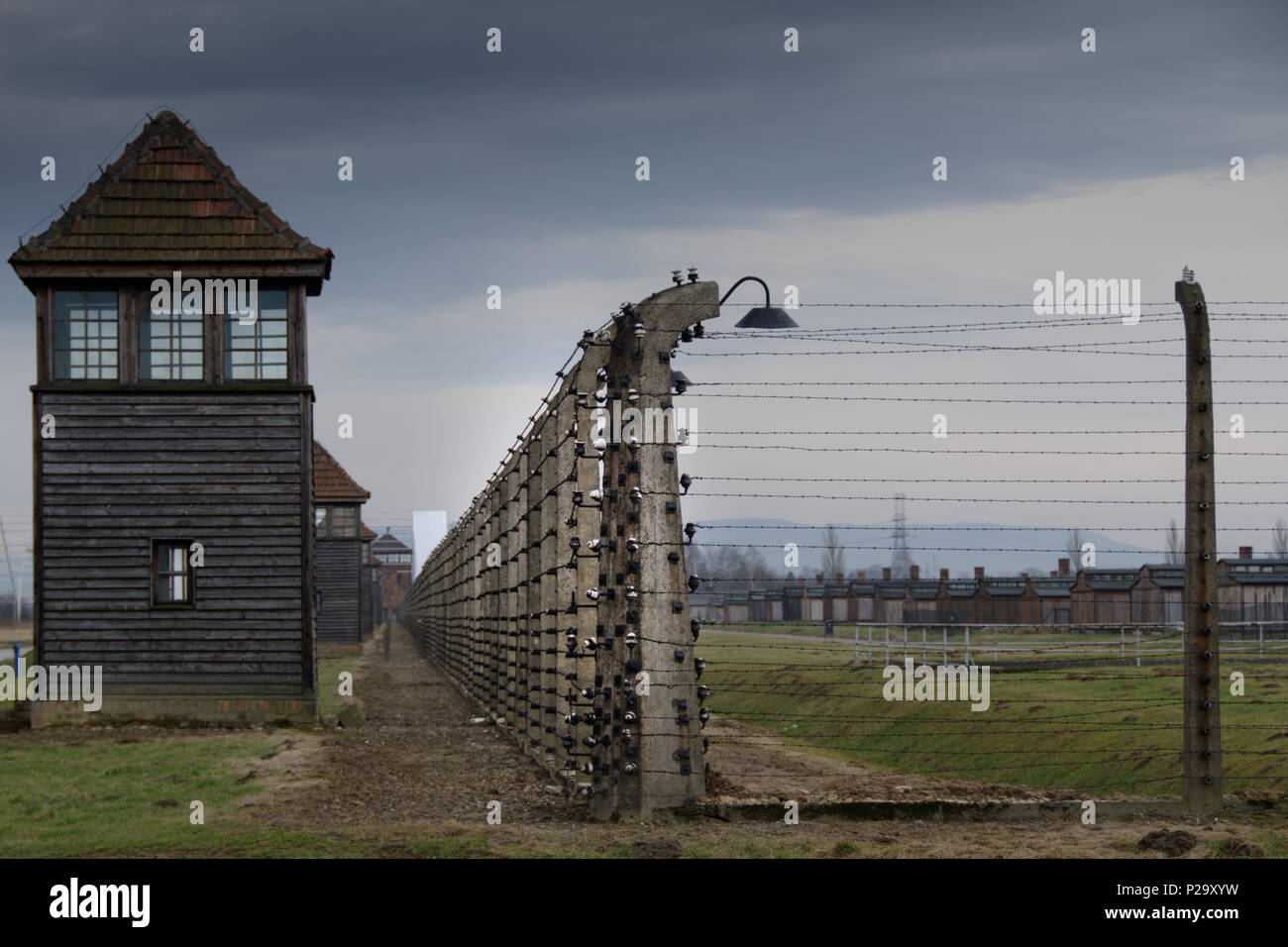 Eine Reihe von Wachtürmen sitzt entlang einer elektrifizierten Zaunlinie in den Ruinen von Auschwitz-Birkenau mit Kaminstapeln im Hintergrund. Stockfoto