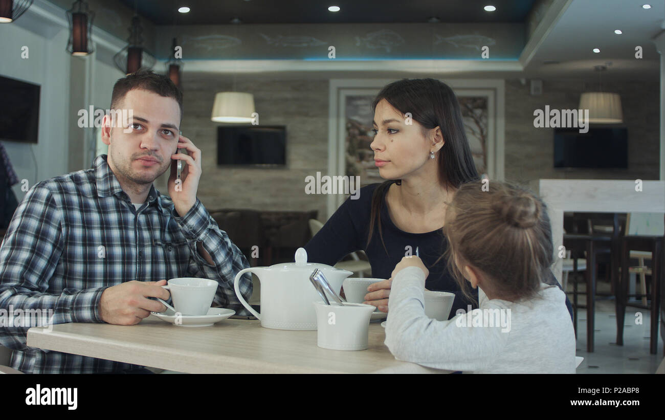 Junge Familie im Cafe. Vater einen Anruf tätigen, während Mutter und Tochter sprechen. Mutter auf ihn schaut unzufrieden. Stockfoto