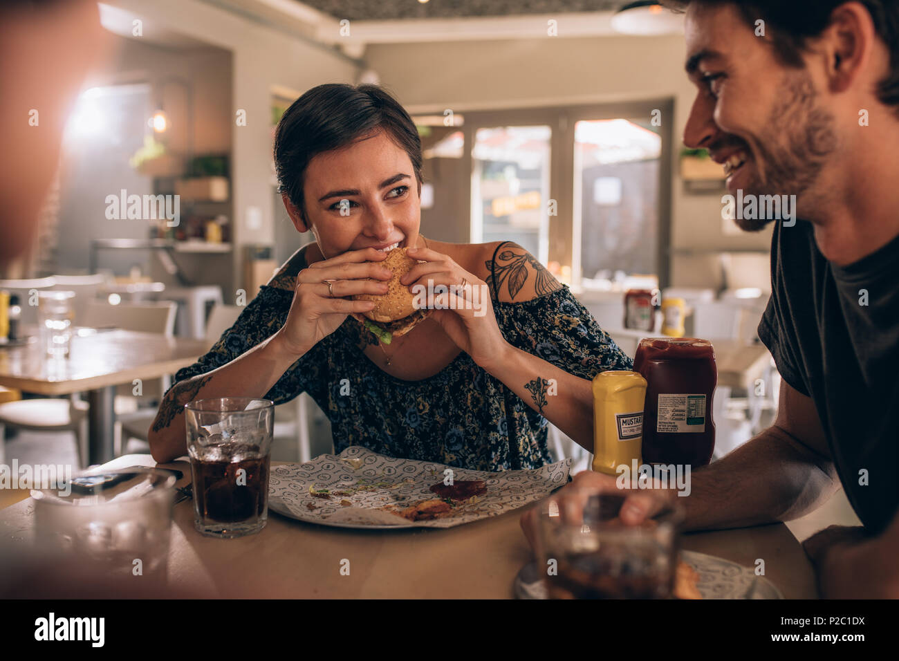 Junge Frau essen Burger beim Sitzen mit Freunden im Restaurant. Junge Leute plaudern und Burger essen im Cafe. Stockfoto
