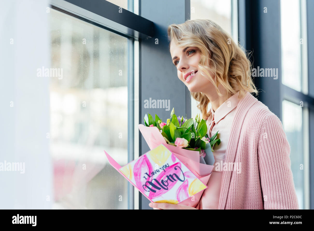 Lächelnde Frau mit Blumenstrauß und Ich liebe dich Mama Postkarte in den Händen, Fenster, Muttertag Konzept Stockfoto