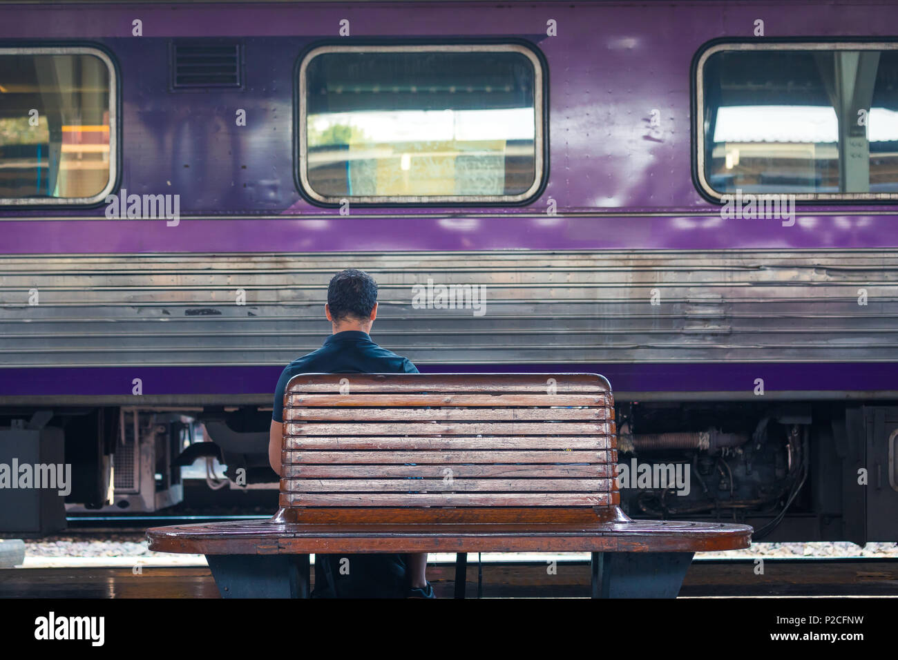 Zurück von einem Mann auf einer Bank am Bahnhof mit lila Zug wartet im Hintergrund Stockfoto