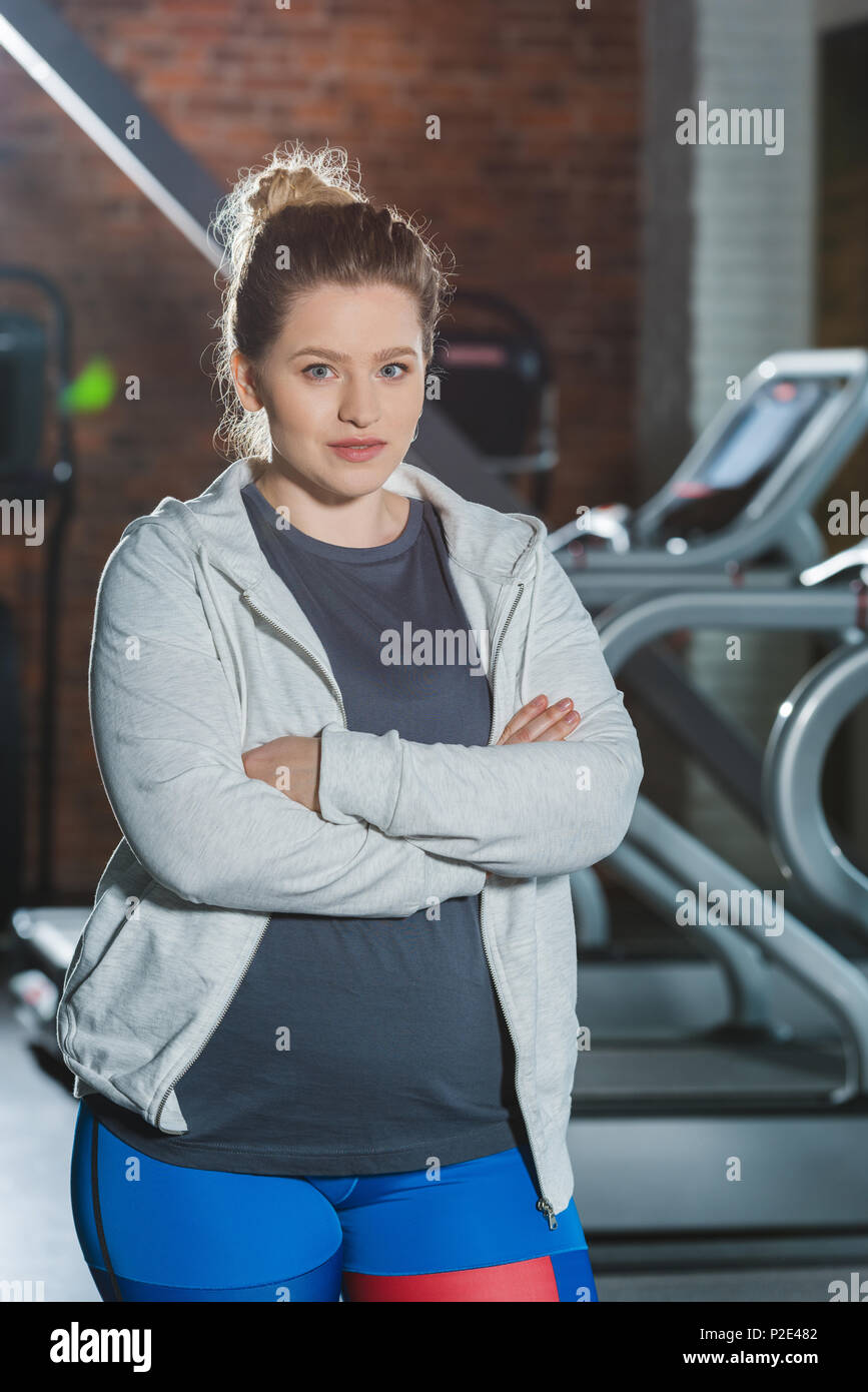 Übergewichtige Frau, die im Fitnessstudio mit verschränkten Armen und Kamera Stockfoto