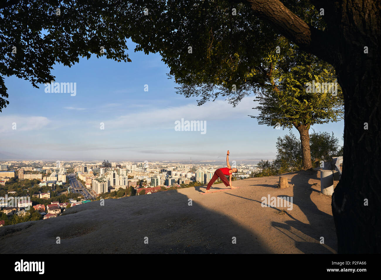 Passen Mann im roten Kostüm Yoga Asana Dreieck im Park mit die Stadt und die Berge im Hintergrund in Almaty, Kasachstan Stockfoto
