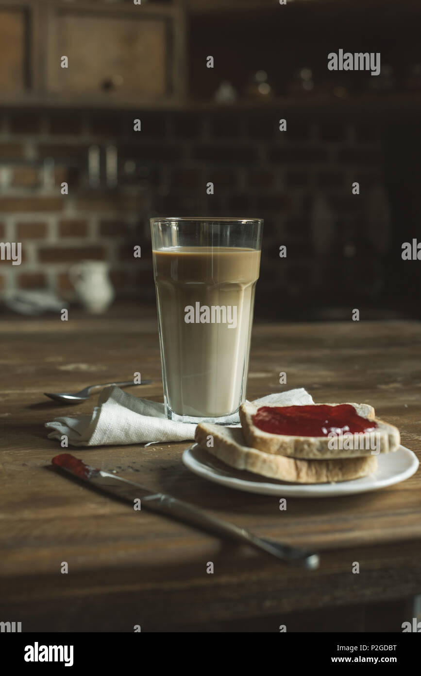 Latte in Glas, Brot mit Marmelade auf dem Teller und Besteck auf hölzernen Tischplatte Stockfoto