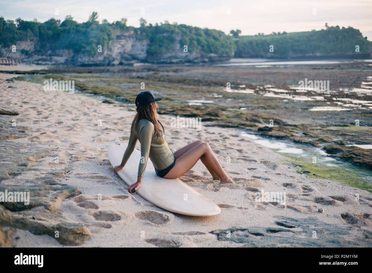 Junge attraktive Frau im Neoprenanzug sitzen auf Surfboard auf Küste und suchen bei Sonnenuntergang Stockfoto