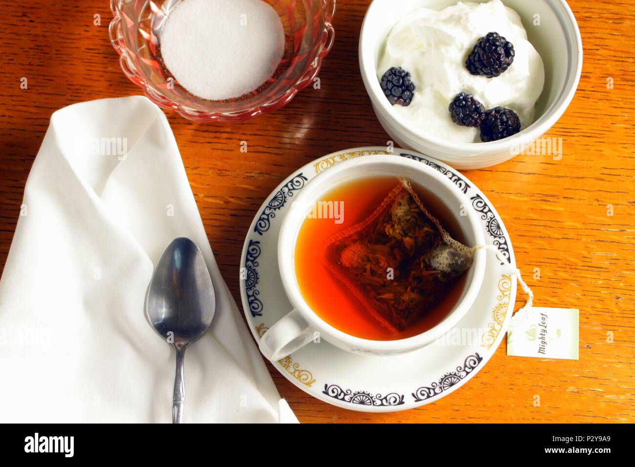 Teetasse und Pudding Dessert mit schwarzen Beeren und einem Tuch Serviette sitzen auf einem Holz Couchtisch mit einem Löffel und Zucker. Stockfoto
