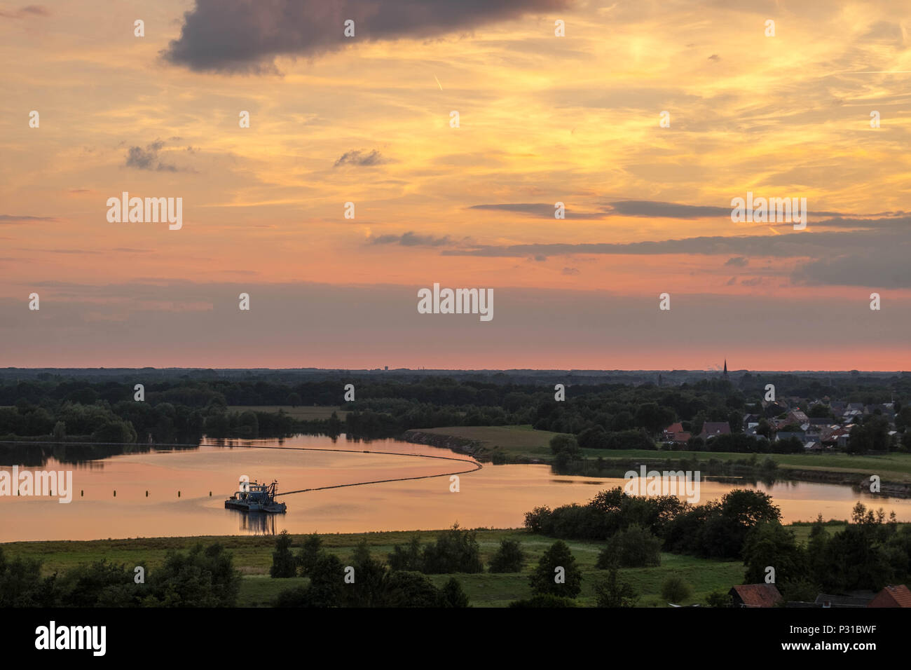 Eines schwimmbaggers liegt auf einem Teich in der Landschaft der Region "Kempen" in Belgien. Auf der rechten Seite sehen Sie die Häuser der Stadt "Essel". Die Sonne untergeht. Stockfoto