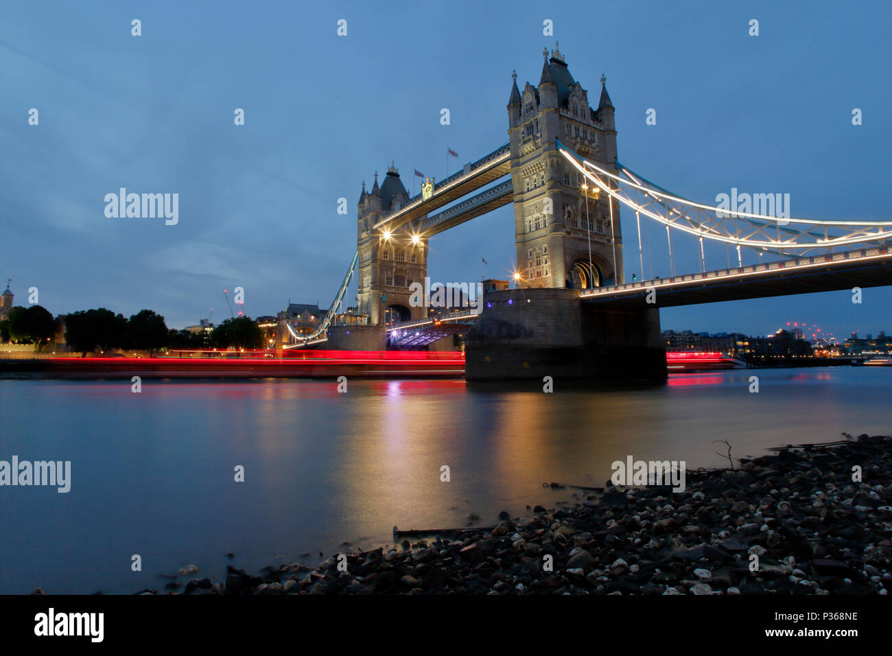 Die Tower Bridge, von einer niedrigen POV bei Ebbe auf der Themse Vorland, eine leichte Spur von einem vorbeifahrenden Party Boot leuchtet rot und reflektiert in der Themse Stockfoto
