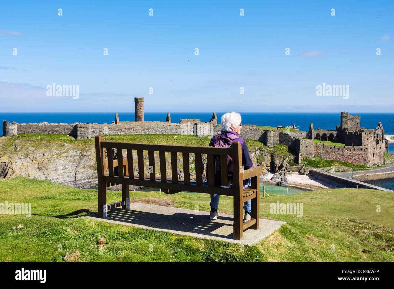 Ältere Rentnerin OAP Dame entspannen allein im Urlaub sitzen auf einer Bank auf dem Hügel mit Blick auf Peel Burgruinen. Peel, Isle of man, Britische Inseln Stockfoto