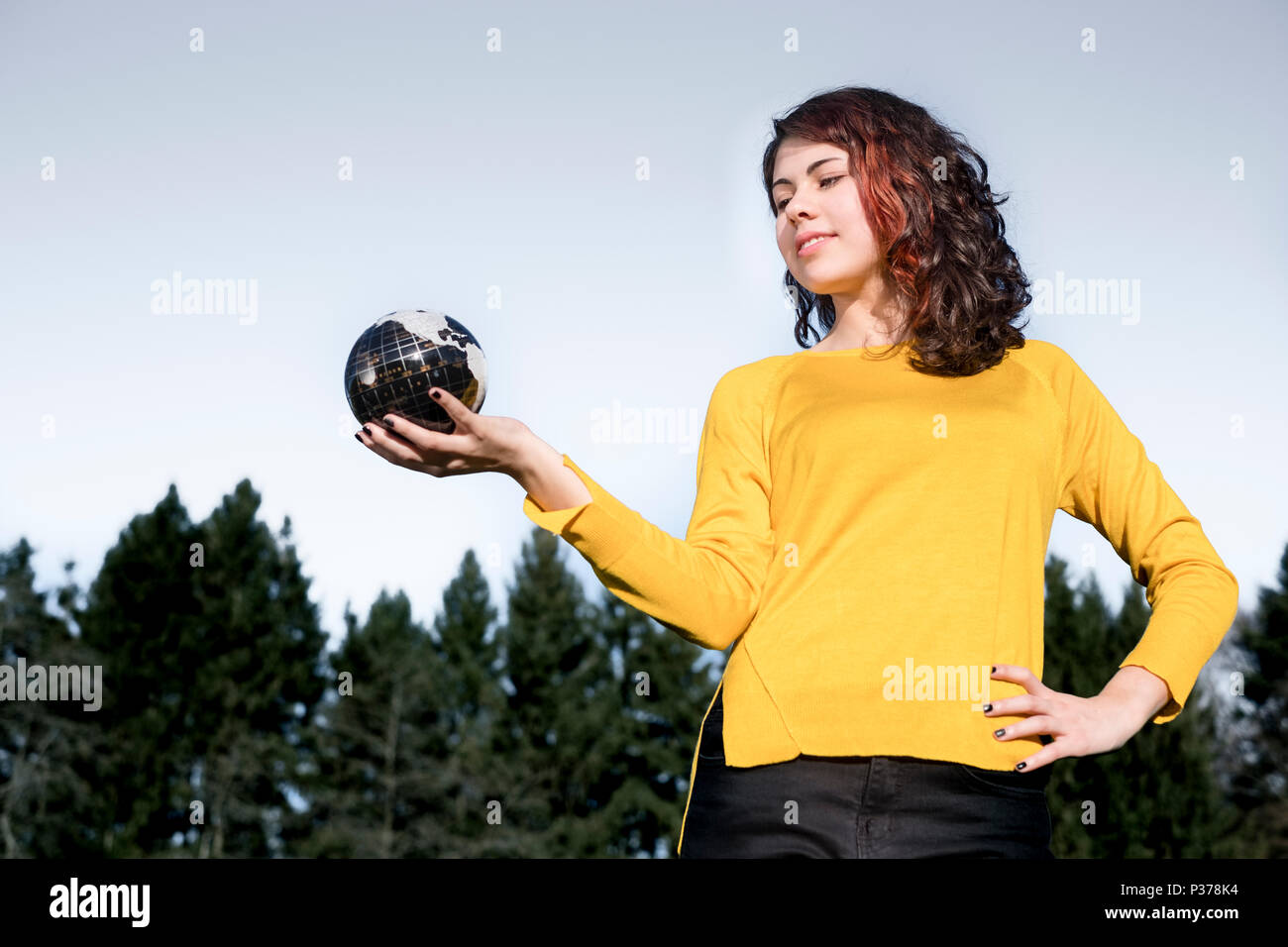 Die ganze Welt in Ihrer Hand: junge Frau in stilvollen gelb Pullover holding Erde in ihre Hand vor Himmel und Bäumen. Speichern Sie die Erde Konzept. Stockfoto