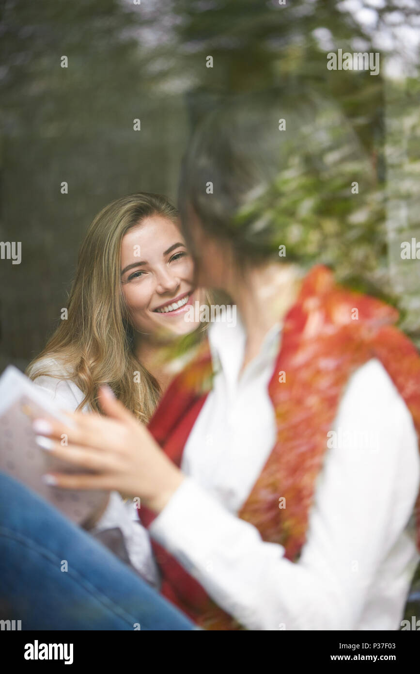 Hübsches Mädchen lächelnd zu ihr Freund hinter Fensterglas sitzen. Die Frauen, die klassischen, weißen Hemden und Strickjacken, blaue Jeans. Kaffee trinken, eine gute Zeit habend, die ror-Projekt. Stockfoto