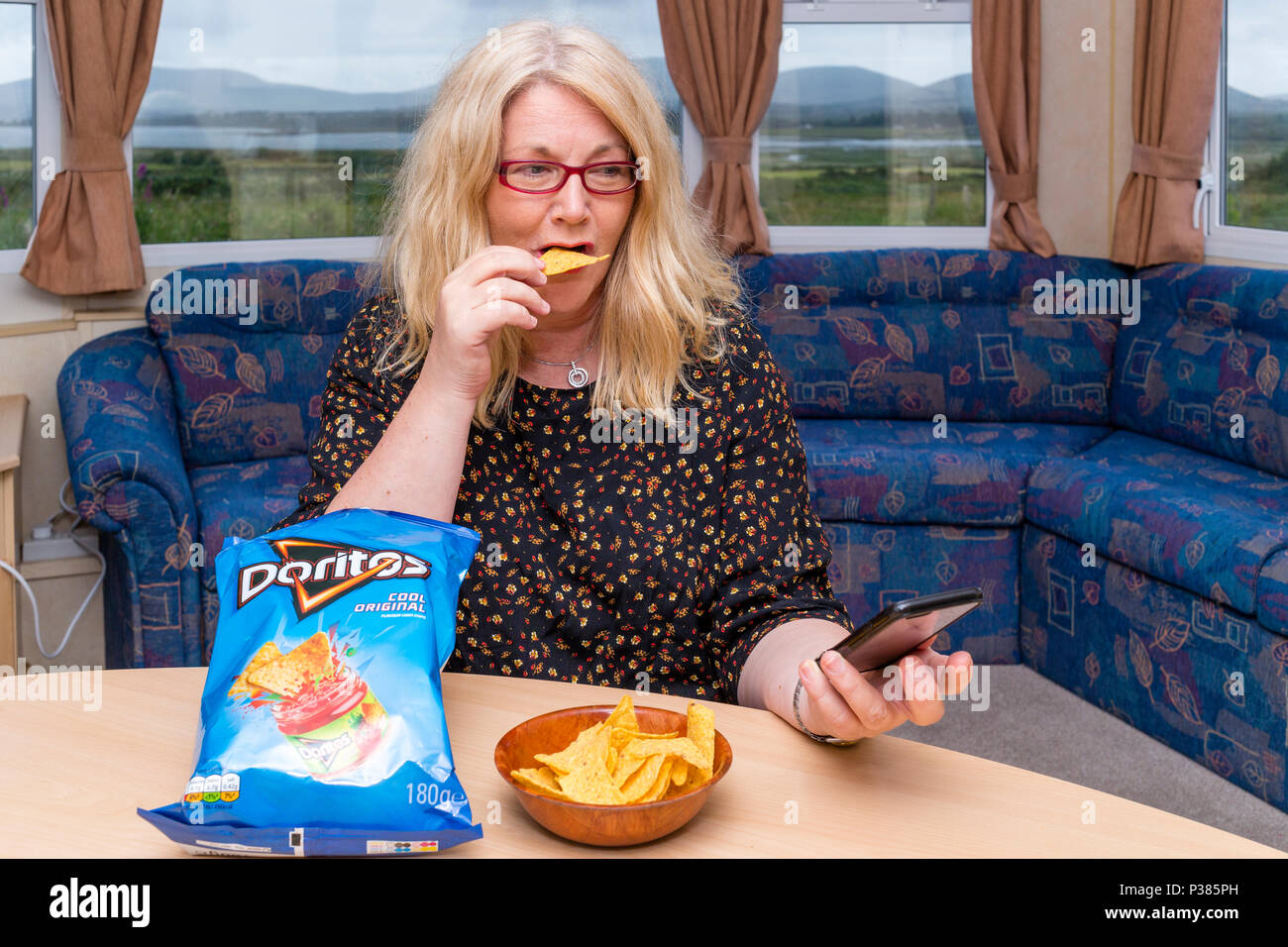 Blonde Frau isst Doritos aus ein Teilen Größe Tasche und eine kleine Schüssel Stockfoto
