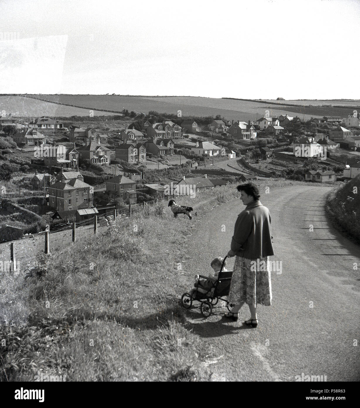 1950er Jahre, eine Dame mit einem Kleinkind Kind im Kinderwagen und Hund - weg von der Leitung - stehend auf einer leeren Straße auf einem Hügel mit Blick auf eine Stadt, England, UK. Stockfoto