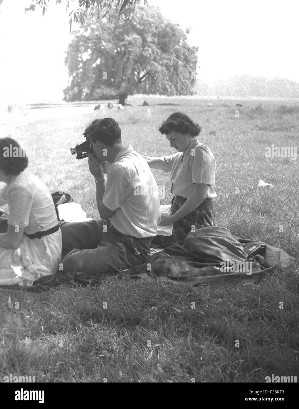 1950er Jahre, historische Bild, Sitzen im Freien auf einer Wolldecke auf einem grasbewachsenen Ufer in einer Parklandschaft mit zwei Damen, ein Herr mit einer Filmkamera, England, UK. Stockfoto