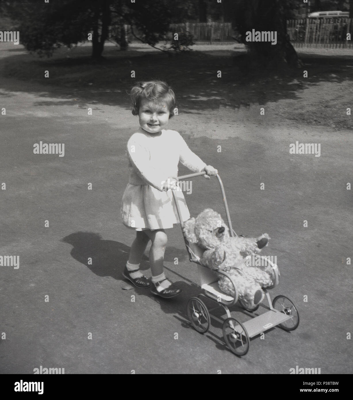 1950, historische, junges Mädchen, im Freien in einem Park schieben ihr Spielzeug metall Kinderwagen mit Teddybär sitzen, entlang eines Pfades, England, UK. Stockfoto