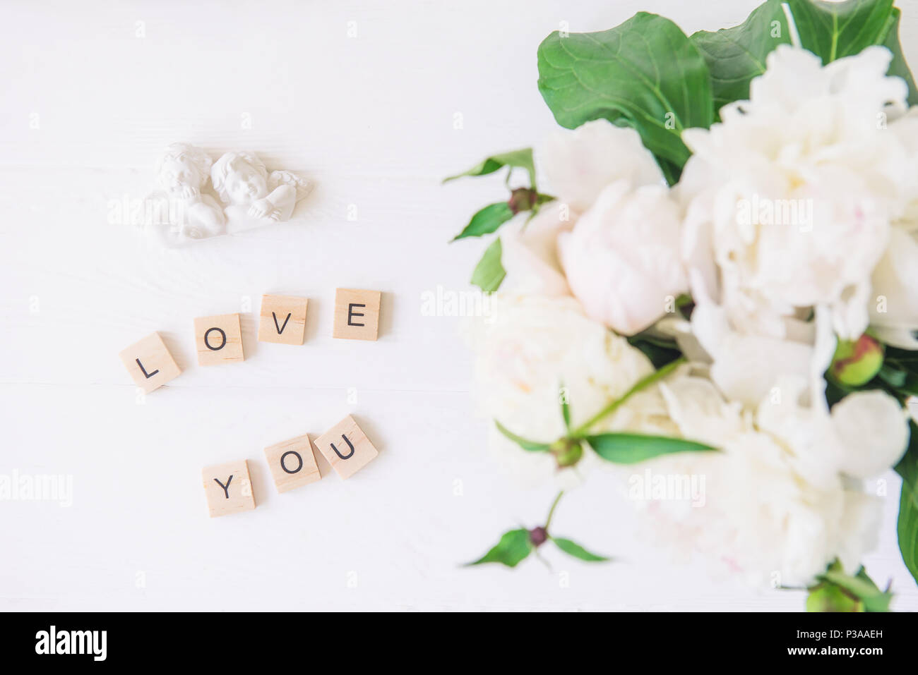 Flach Liebe dich Schriftzug in Holzblöcke mit statuette von zwei antike wunderschöne Engel mit unscharfen weiße Pfingstrose auf hellen Hintergrund geschrieben. Liebe Stockfoto