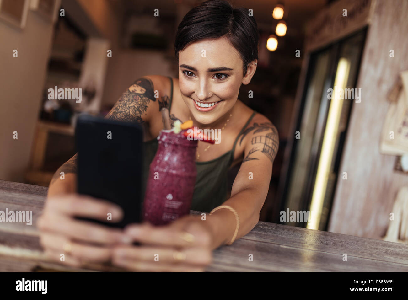 Lächelnde Frau mit einem selfie mit einem Smoothie vor ihr Handy für ihr Essen blog platziert. Essen blogger Fotografieren für Ihren Blog. Stockfoto