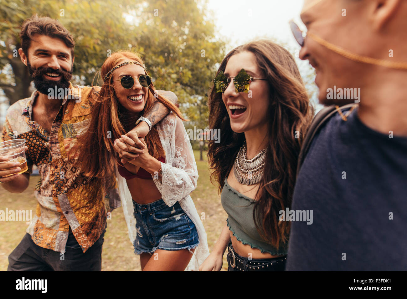 Zwei Paare wandern und lächelnd in einem Park mit Getränken. Junge Menschen im Summer Music Festival. Stockfoto
