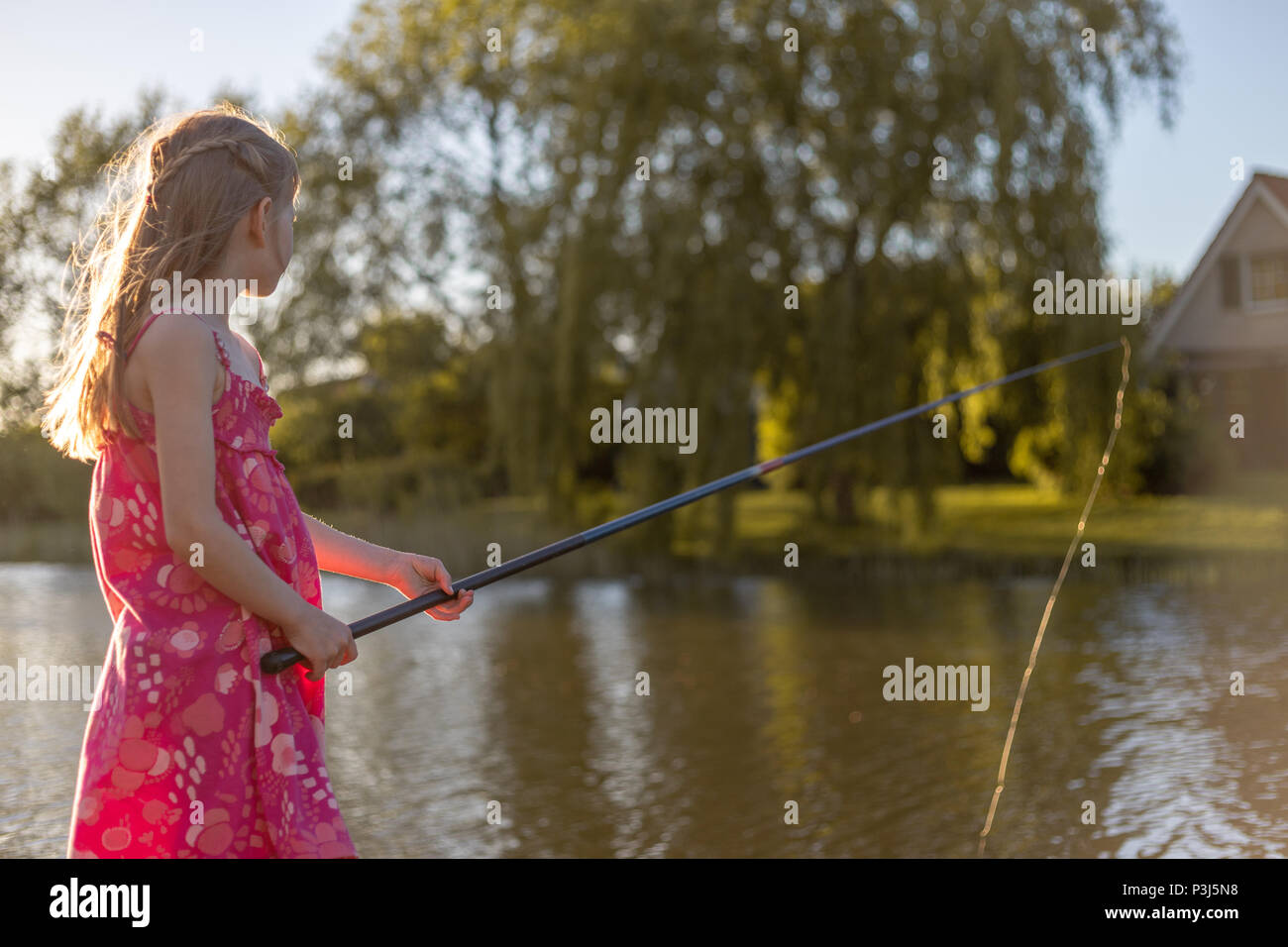 Ein 7 Jahre altes Mädchen steht an einem See mit Angelrute und Blick auf das Wasser. Sie wartet auf einen Fisch zu beißen Stockfoto