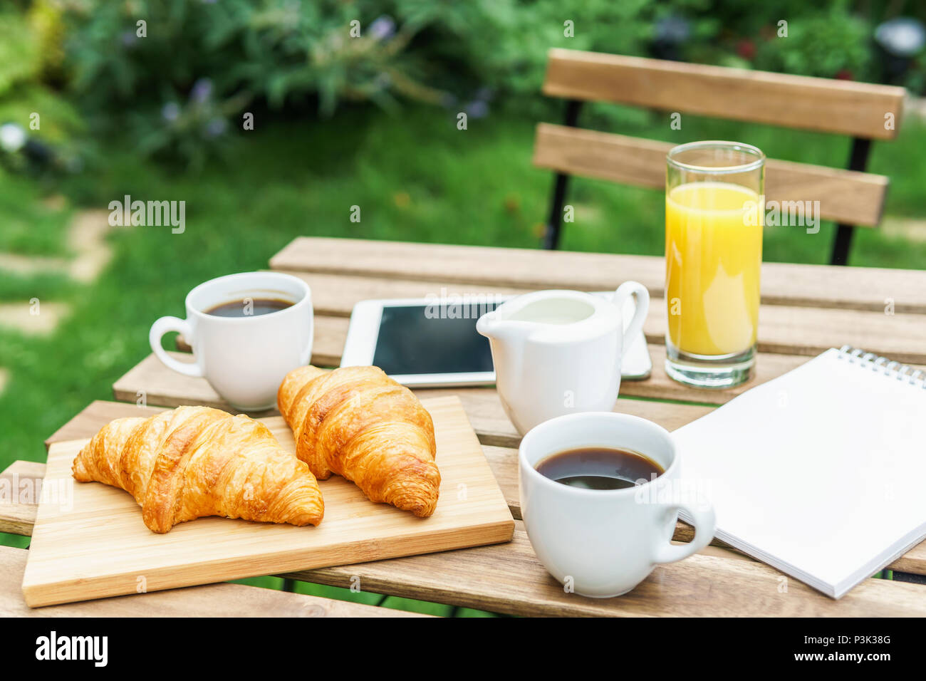 Frühstück im grünen Garten mit französischen Croissants, Kaffee, Orangensaft, Tablet und Hinweise Buch auf hölzernen Tisch Stockfoto