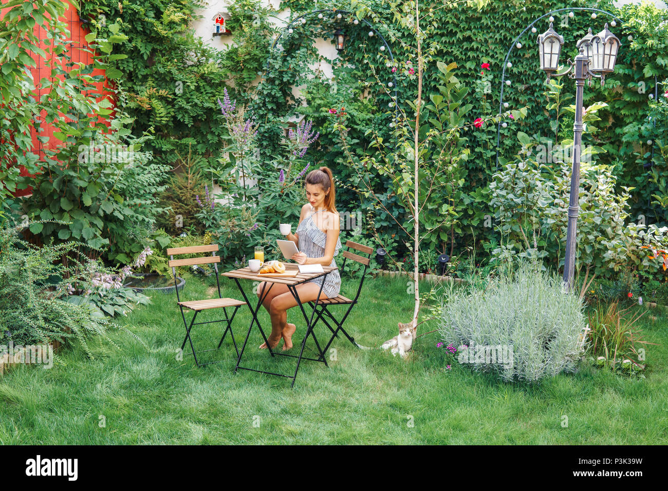 Junge und attraktive Frau mit Frühstück im grünen Garten mit französischen Croissants, Donuts, Kaffee, Orangensaft, Tablet und Hinweise Buch über Stockfoto