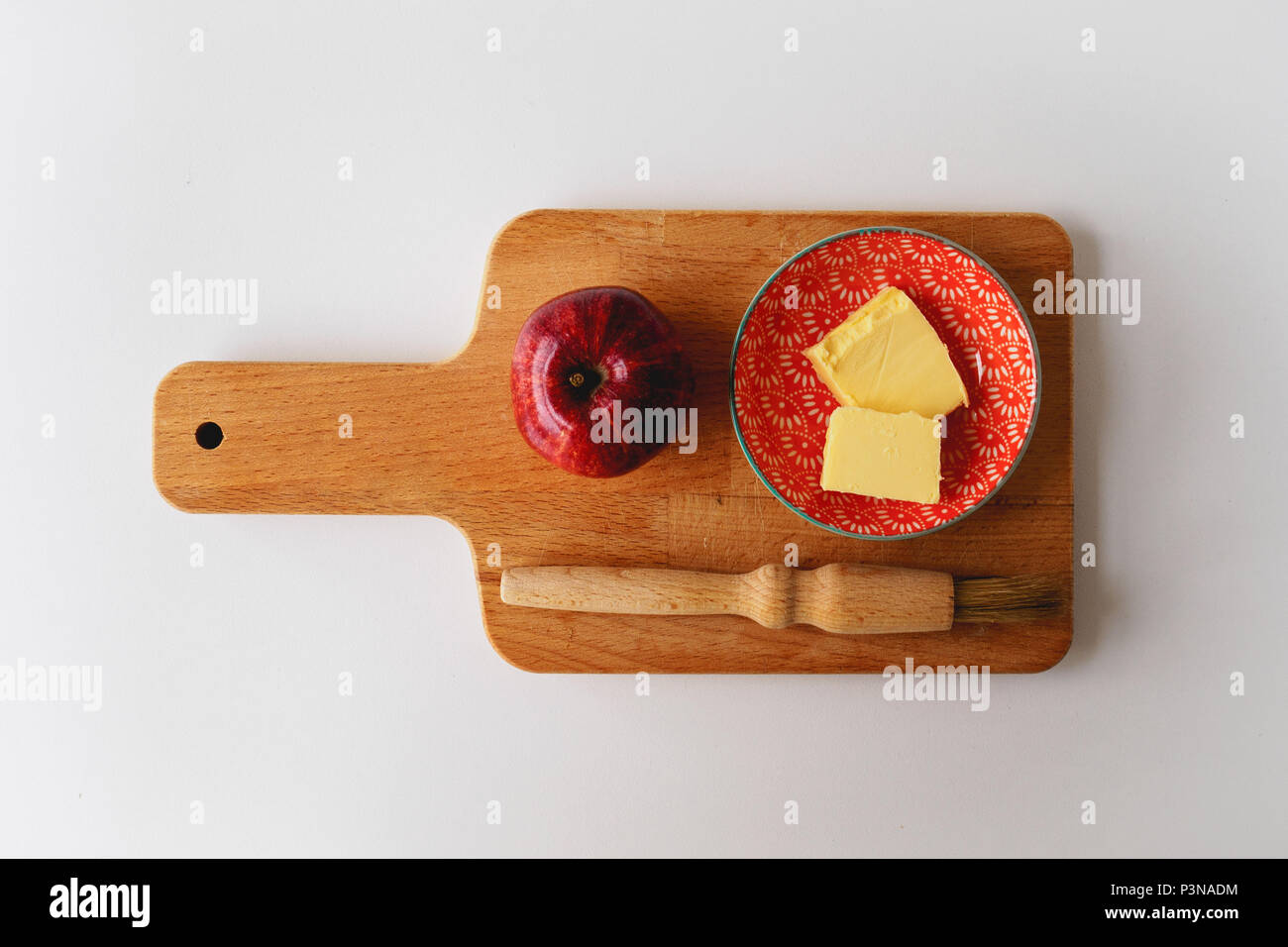 Ein roter Apfel, eine kleine Platte mit Butter und ein backblech Pinsel auf einer hölzernen Schneidebrett mit einem weißen Tag. Zutaten für einen Apfelkuchen. Essen Konzept. Stockfoto