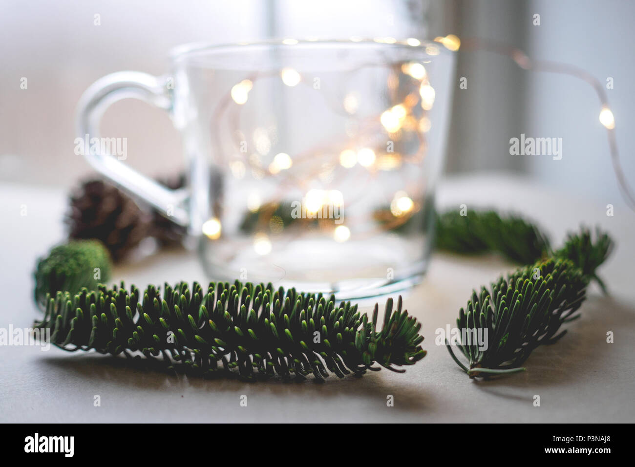 Nahaufnahme einer unscharfen Glas Becher mit warmen Weihnachtsbeleuchtung innen durch Tannenzapfen und Zweige auf einen weißen Tisch umgeben. Weihnachten Konzept. Stockfoto