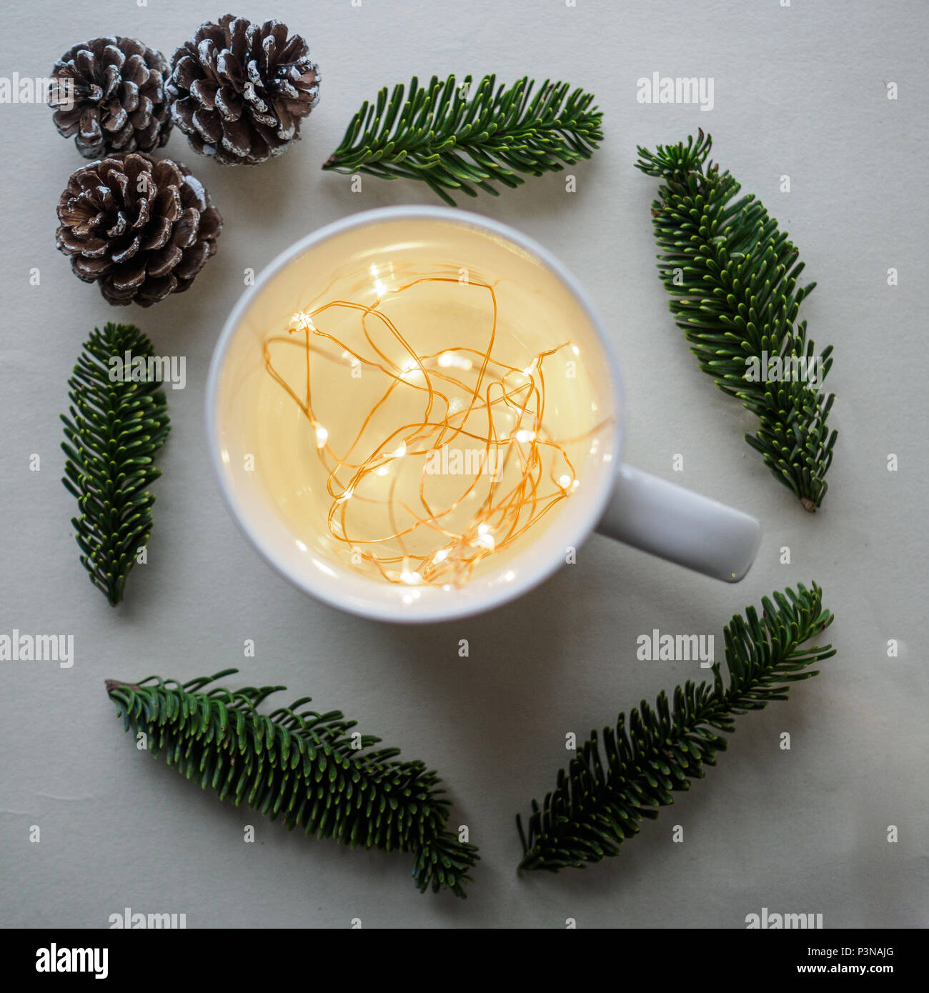Blick von oben auf eine weiße Tasse mit warmen Weihnachtsbeleuchtung innen durch Tannenzapfen und Zweige auf einen weißen Tisch umgeben. Weihnachten Konzept. Quadratischen Format. Stockfoto