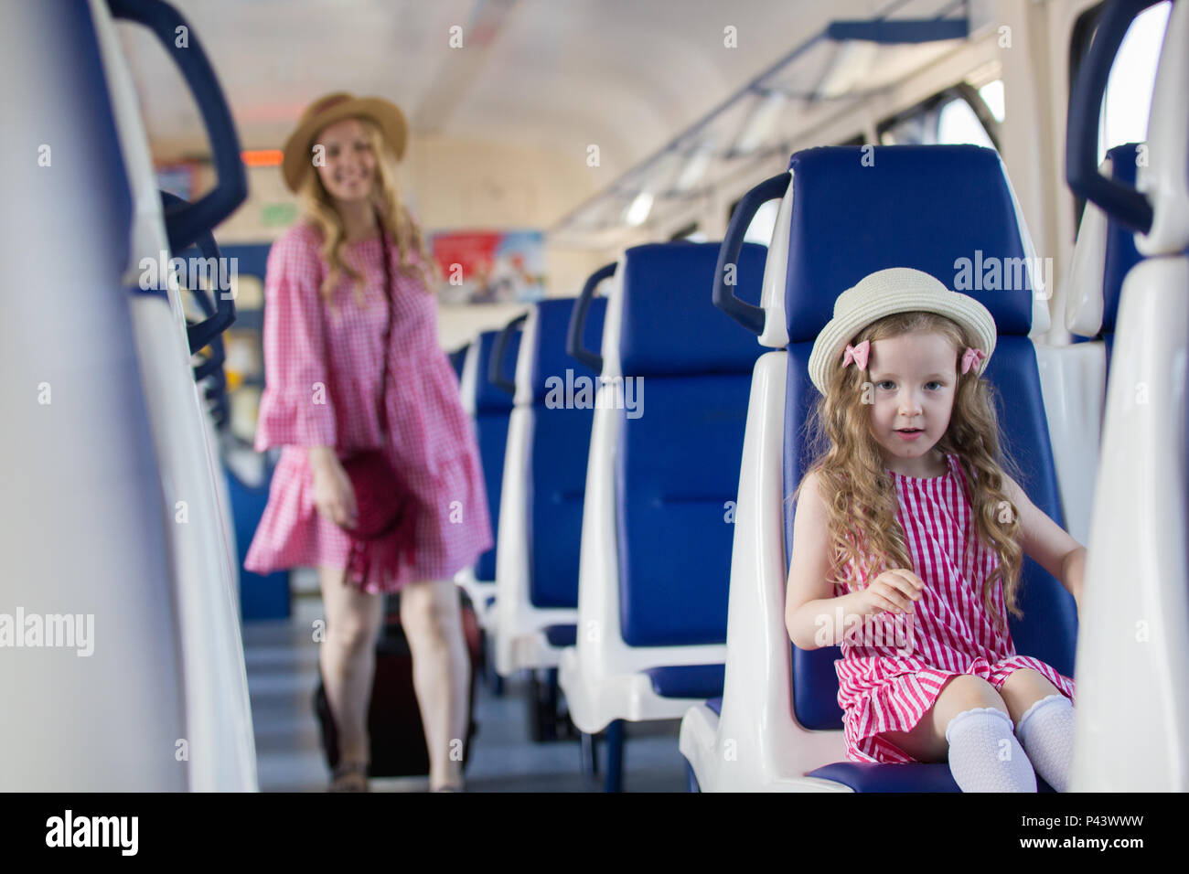 Glückliche junge Mutter geht mit Koffer zu ihrer kleinen Tochter, die auf Sessel im Zug sitzen Stockfoto