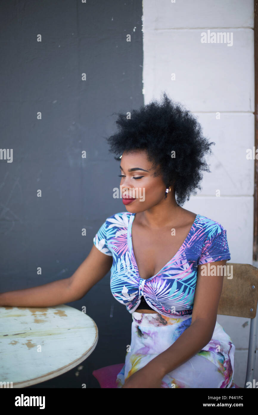 Ein afrikanisches Modell wirft, während an einem Tisch in einem floralen Crop Top gegen eine dunkel kontrastiert Wand saß Stockfoto