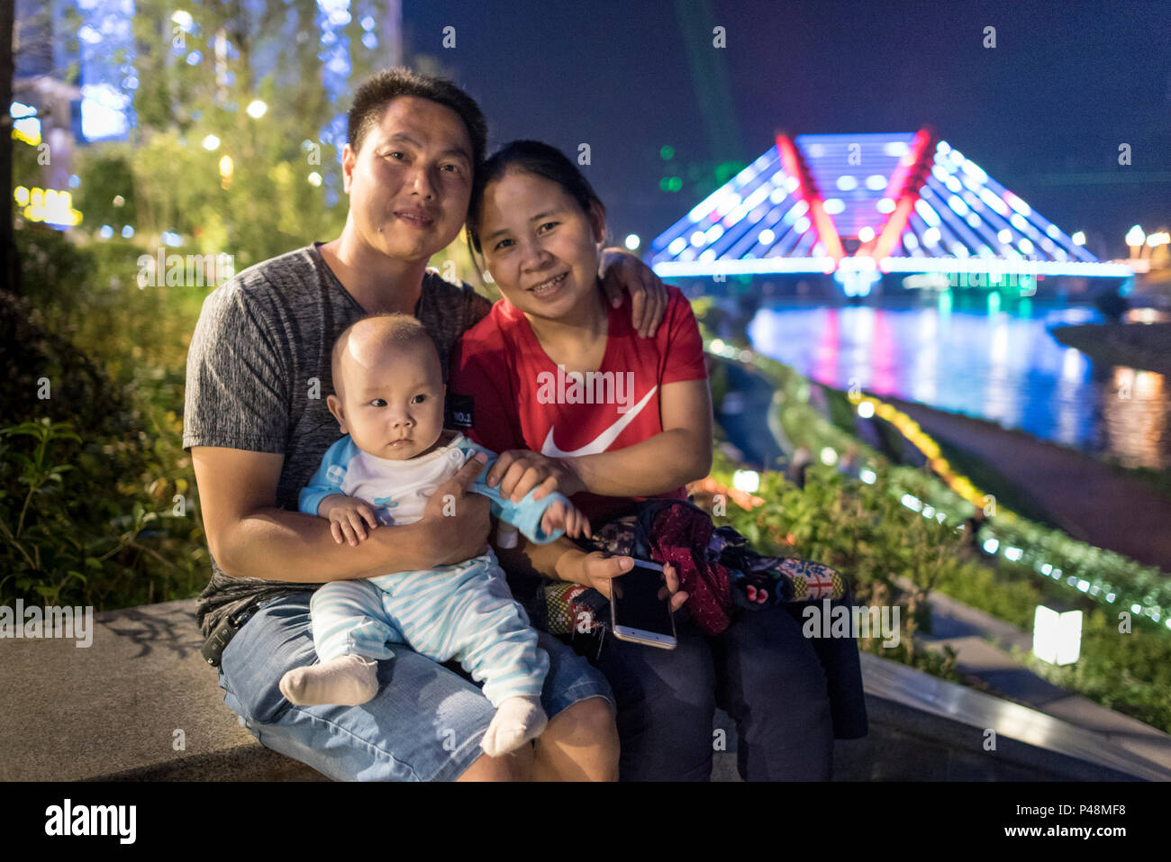 Mutter und Vater mit Baby am Ufer des Flusses und Neon beleuchtete Brücke, nachts das Leben auf der Straße, Libo, Provinz Guizhou, China. Stockfoto