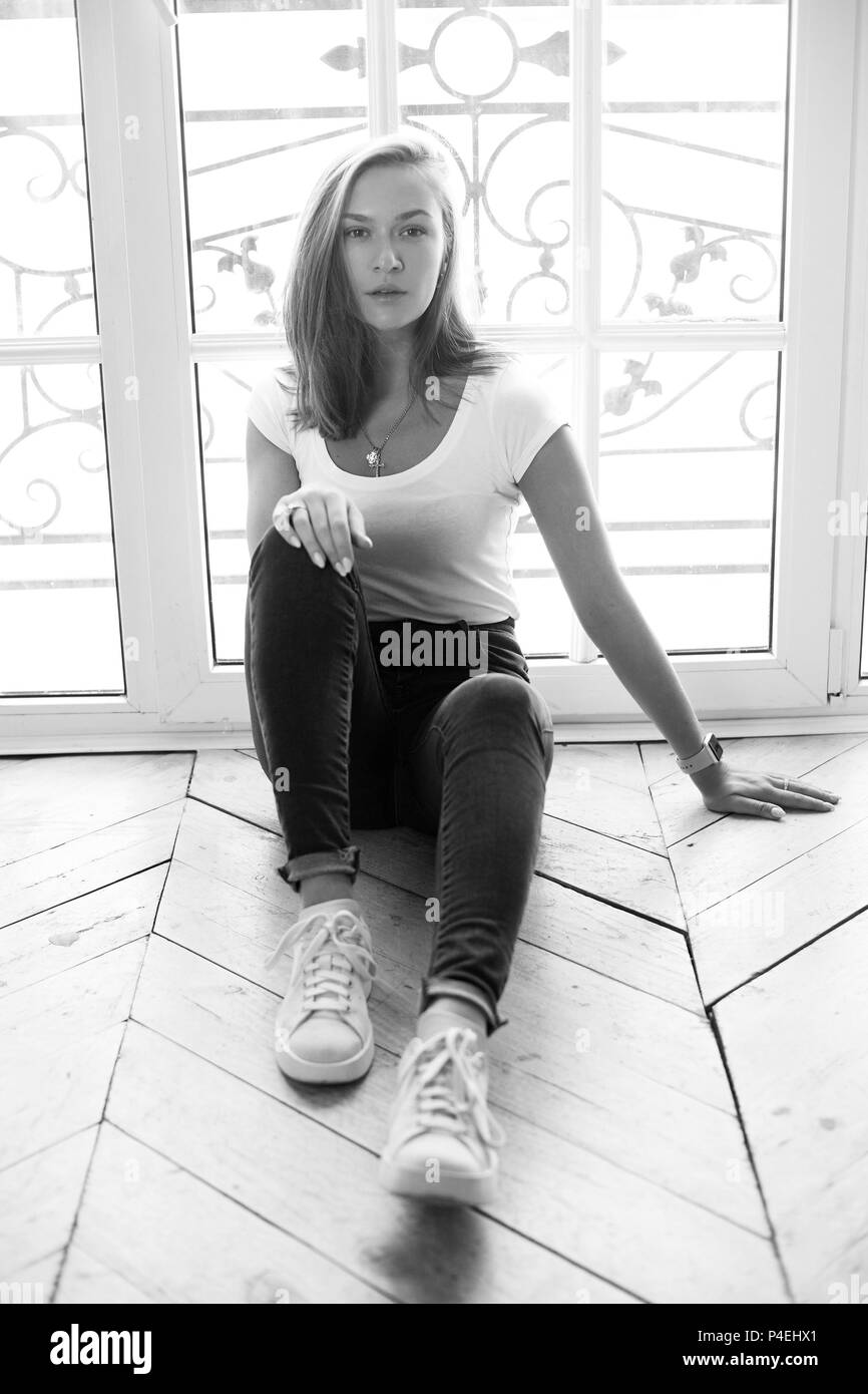 Junge elegante Frau mit langen, geraden Haar zu tragen weißes T-Shirt und Blue Jeans sitzen auf dem Boden gegen die grossen Fenster und Suchen in der Kamera Stockfoto