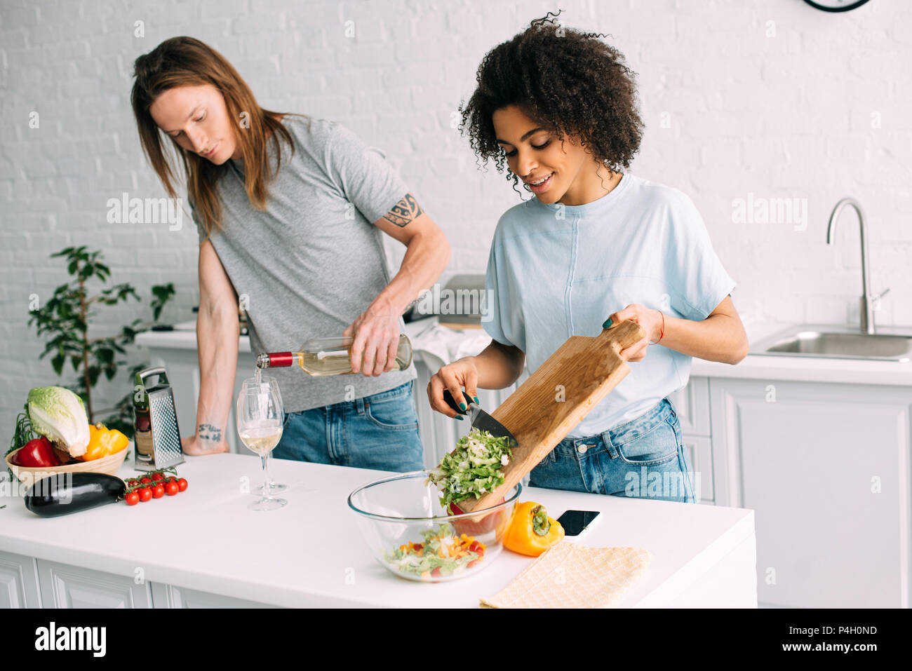 Junge afrikanische amerikanische Frau kochen Salat und Freund gießen Weißwein in Glas Stockfoto