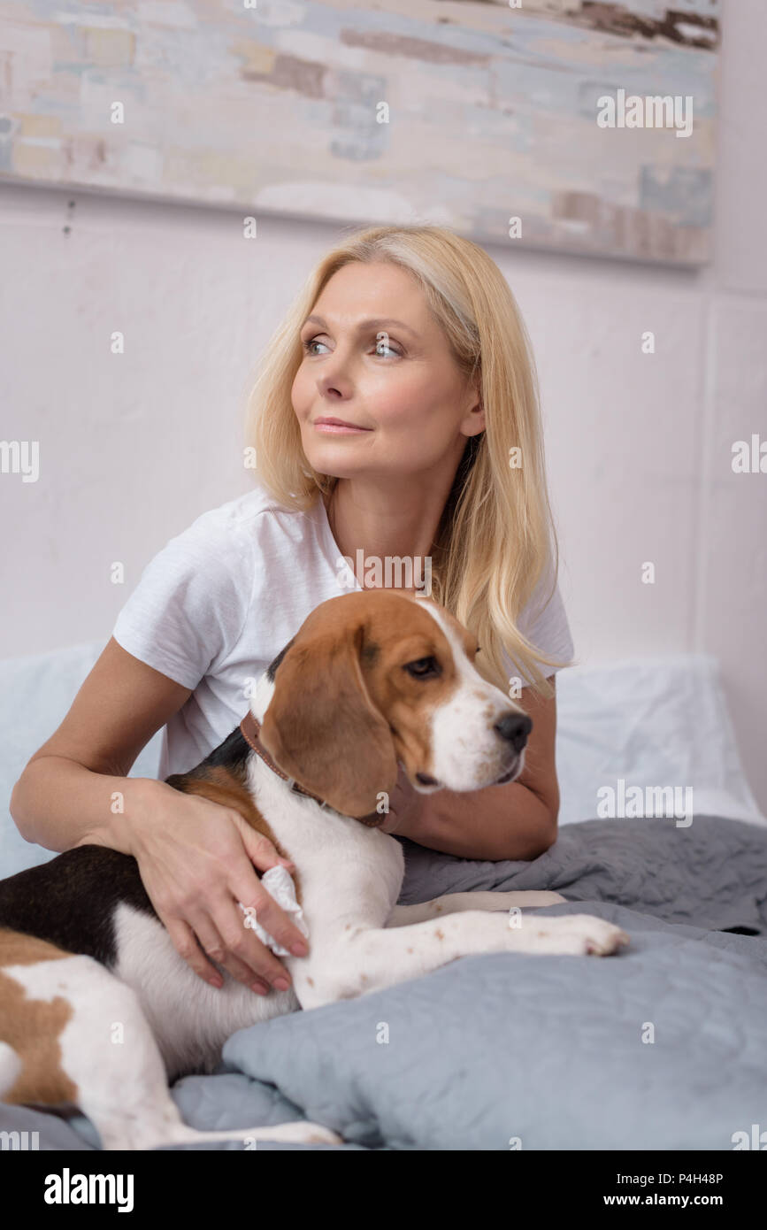 Schöne blonde Frau umarmen Hund und weg schauen beim Sitzen auf dem Bett Stockfoto