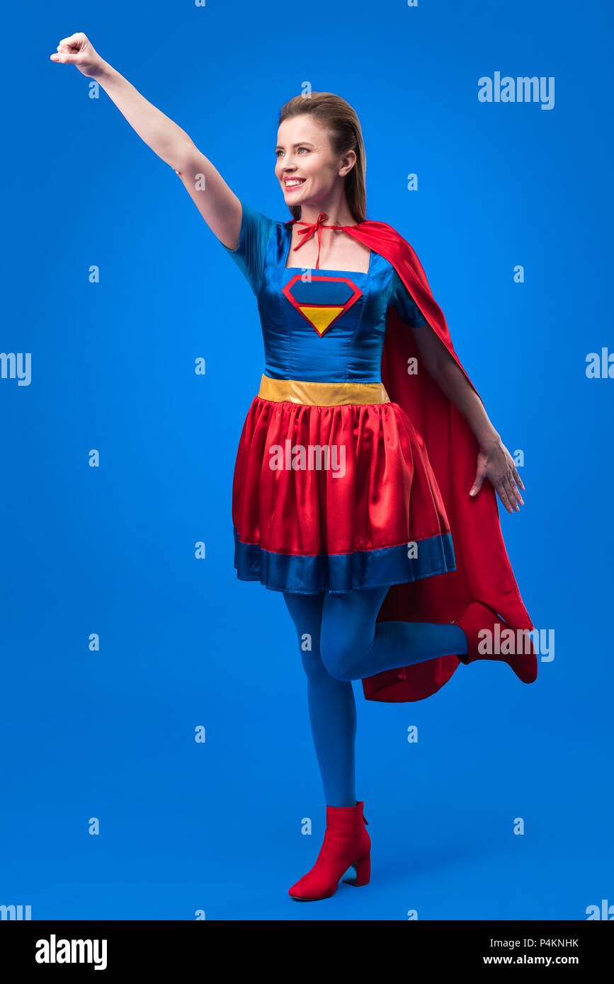 Lächelnde Frau in Superhelden Kostüm mit ausgestrecktem Arm isoliert auf Blau Stockfoto