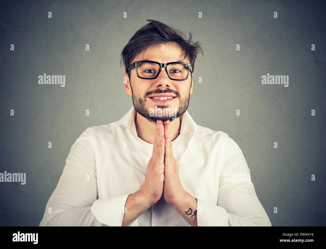 Junge Bartgeier Kerl in Gläsern und Shirt Hände halten zusammen betteln Geste und bitten um Gnade auf grauem Hintergrund Stockfoto