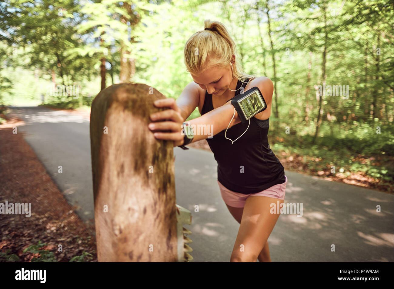 Passen junge blonde Frau in Sportkleidung stützte sich auf eine Stelle neben einem Wald Weg ausdehnen, bevor sie für einen Lauf Stockfoto