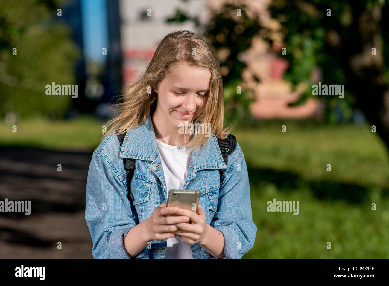 Kleines Mädchen ist blond. Schulmädchen im Sommer nach der Schule. In seinen Händen hält ein Smartphone. Lächelt glücklich. Schreibt Nachrichten in sozialen Netzwerken. Freudige Ruhe nach dem Institut. Stockfoto