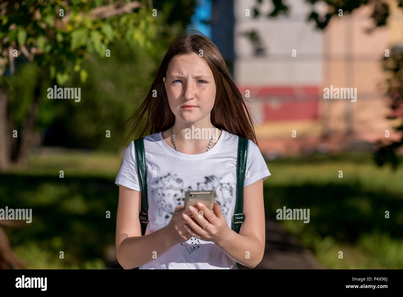 Mädchen Brunette Schulmädchen. Im Sommer in der Stadt in die Natur. In seinen Händen hält ein Smartphone. Das Konzept der ein Anruf an die Eltern. Sieht emotional in den Rahmen. Er Ruhe nach der Schule. Stockfoto