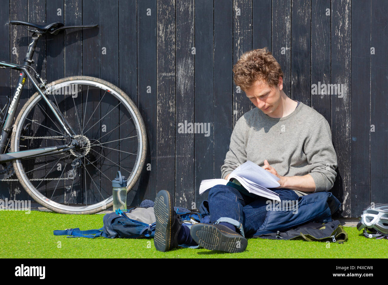 Ein Mann liest ein Buch sitzen auf gefälschte grüne Gras mit einem Fahrrad hielten gegen ein Holz Wand Stockfoto