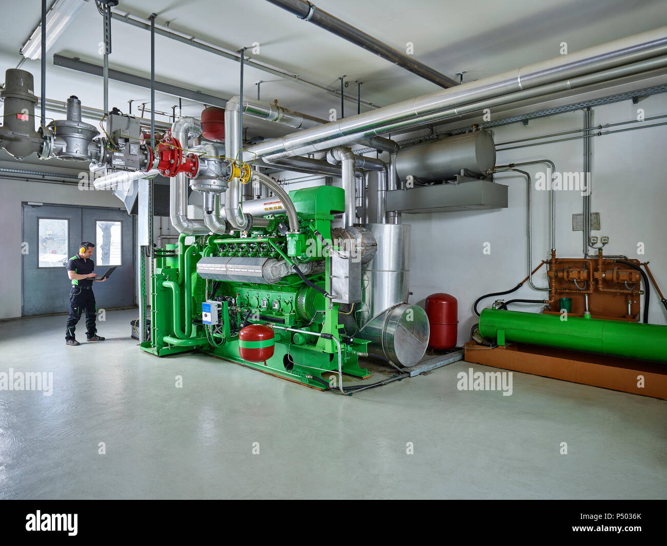 Blockheizkraftwerk, Arbeiter mit Gehörschutz mit Laptop vor Gas Motor Stockfoto