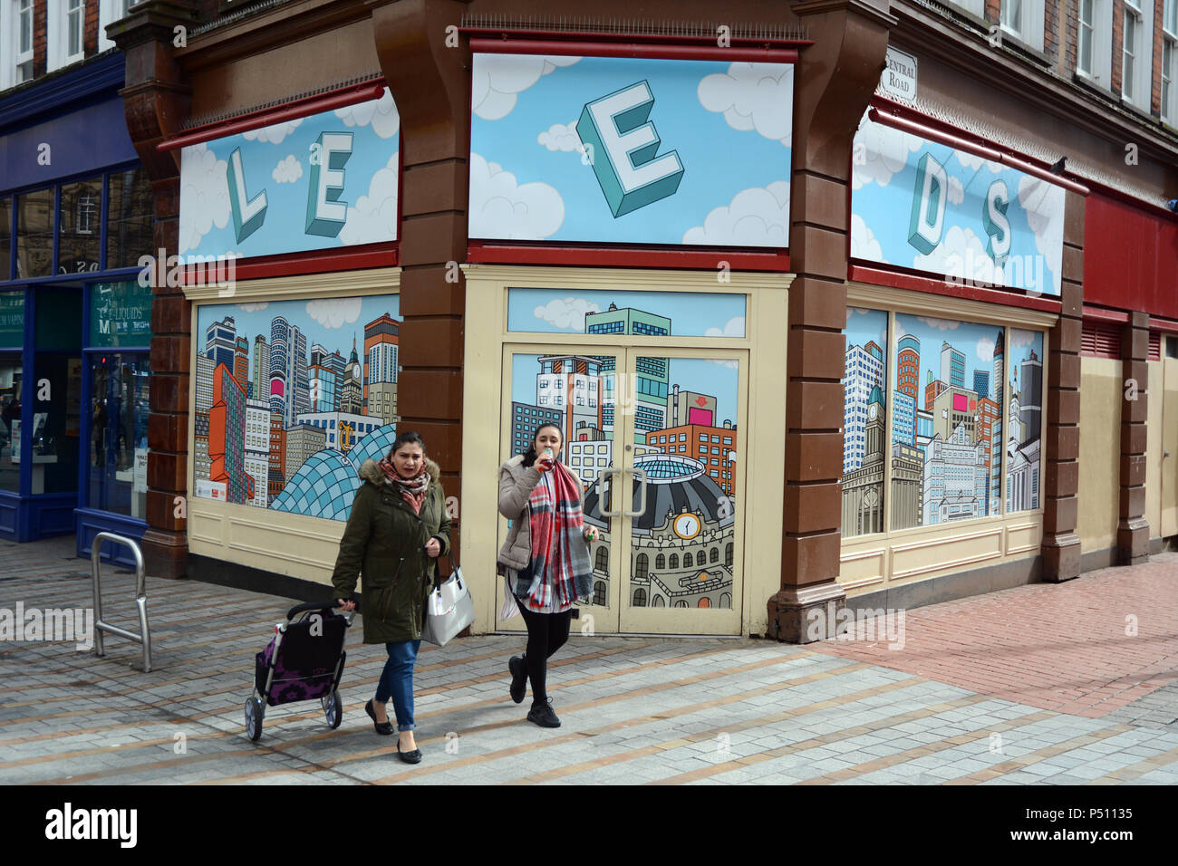 Zwei junge Frauen vorbei an einem leeren Storefront mit einem Hintergrundbild Abbildung der Stadt Leeds, in einer Fußgängerzone, Leeds, England, Vereinigtes Königreich. Stockfoto
