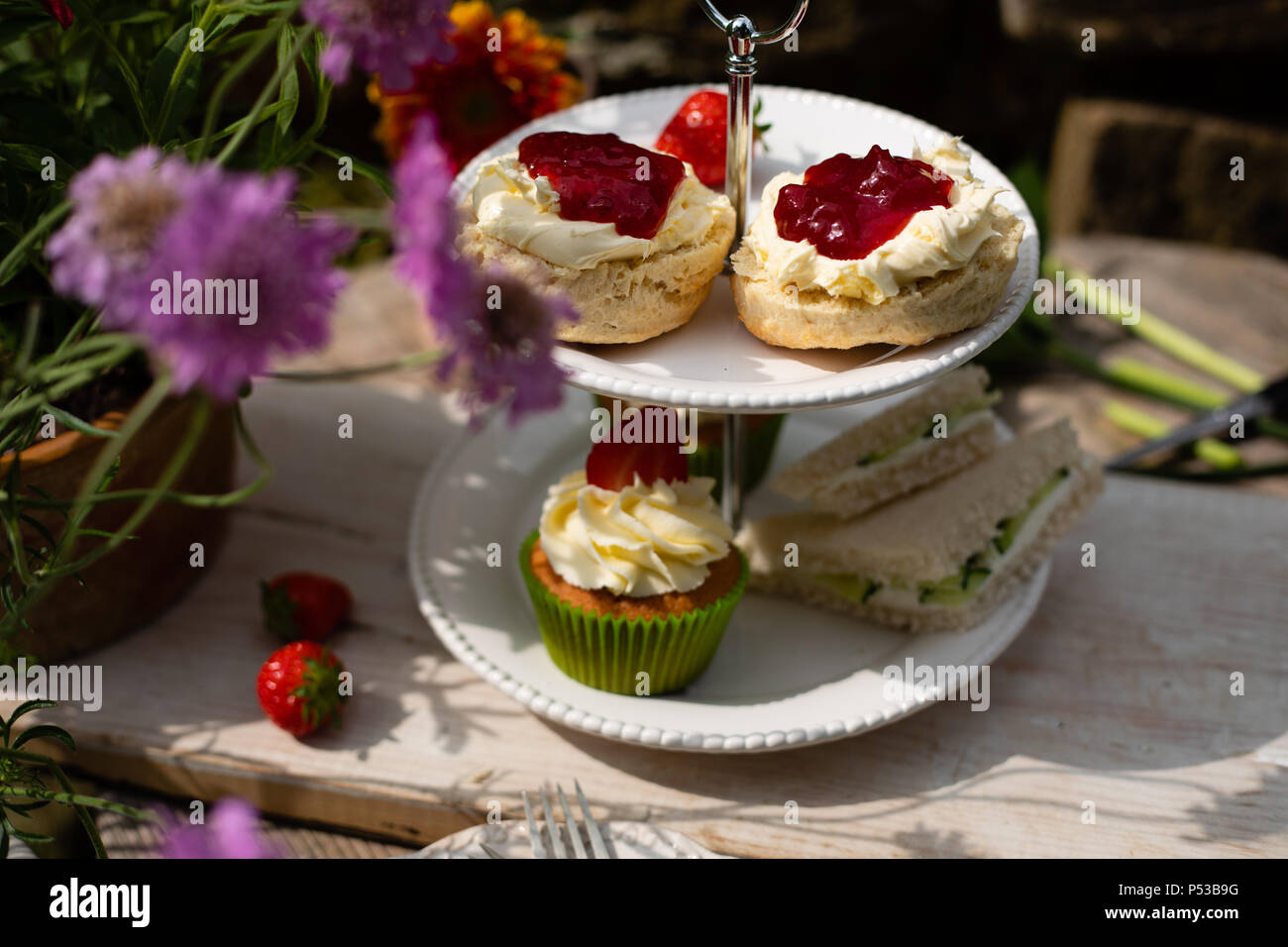 Scones mit Sahne und Marmelade auf Kuchen stand mit Sandwiches und Kuchen im Garten mit Blumen auf Kuchen Stand Stockfoto