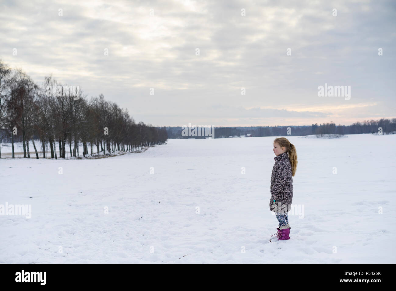 Kleine blonde kaukasischen schwedischen Mädchen stehend Outdoor im Winter Landschaft. Schönen kalten Abend bei Sonnenuntergang mit Schnee auf dem Feld und Allee Stockfoto