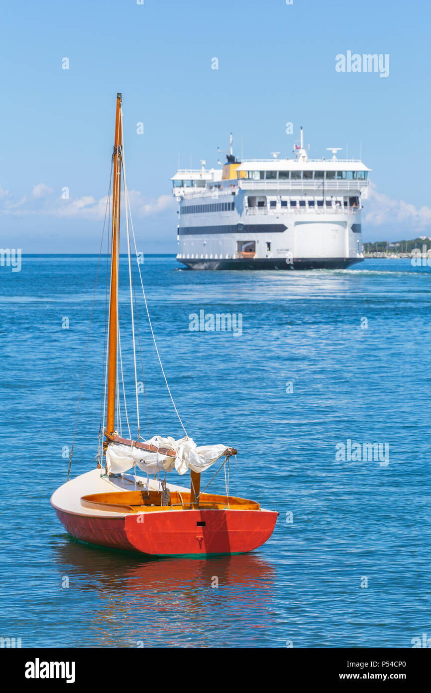 Eine kleine rote hölzerne Segelboot verankert in Küstennähe als Dampfschiff Behörde Fähre Vineyard Haven auf Martha's Vineyard Blätter für das Festland geleitet. Stockfoto