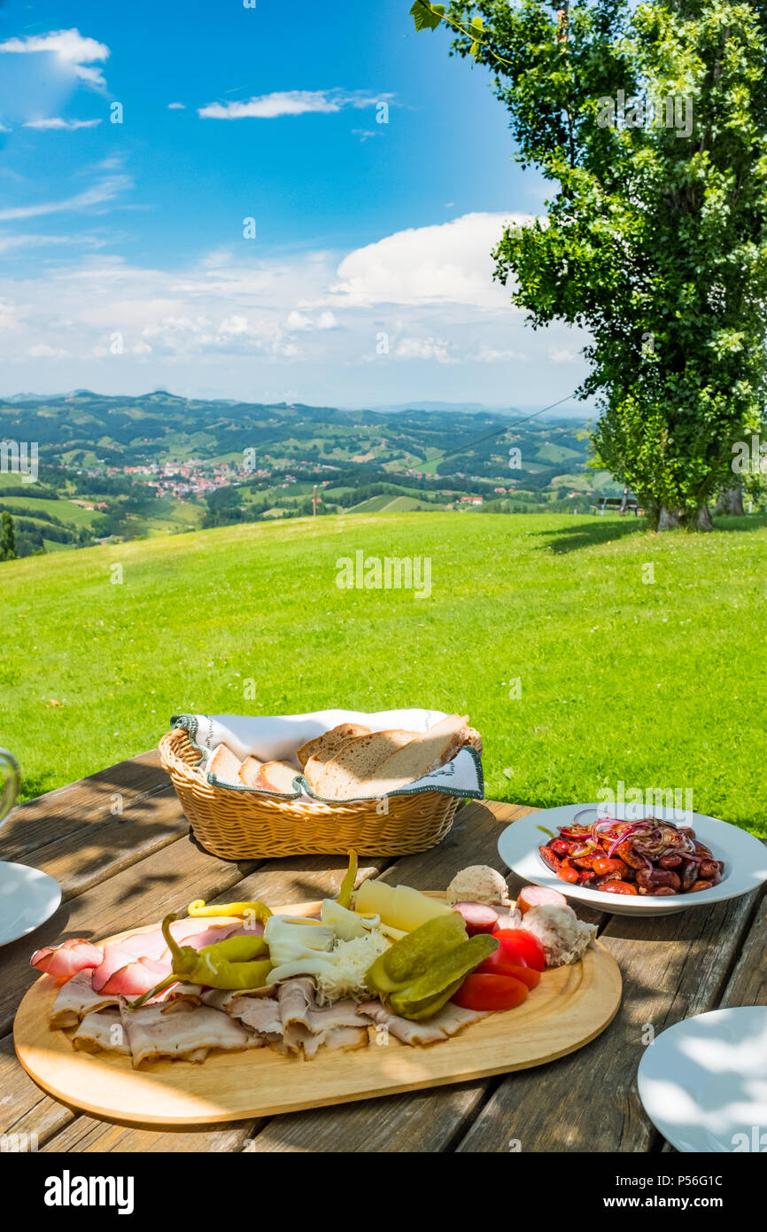 Brettljause mit Aufschnitt, Bohnen, Salat und Brot auf Holztisch mit schönen Blick über grüne Wiese mit Panoramablick auf die südsteirische Weinstraße Stockfoto
