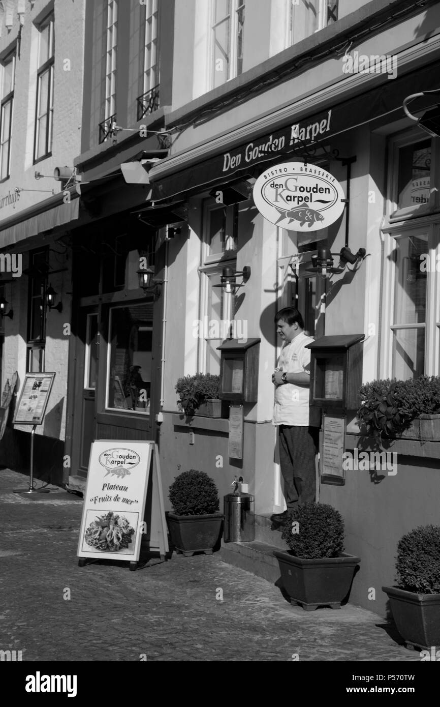 Huidenvettersplein, Brugge, Belgien: ein Kellner steht in der Tür von den Gouden Karpel Restaurant. Schwarz und Weiss Stockfoto